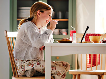 Girl in pajamas with popcorn print, sitting on a wooden chair. Laughing during a morning video call while having breakfast.