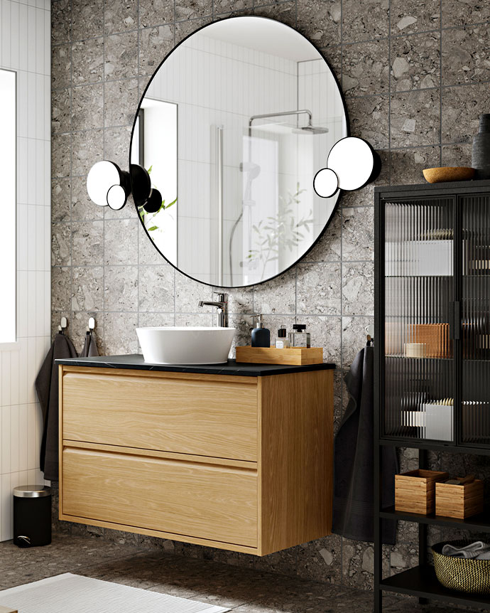 Espejo redondo y tocador flotante de madera clara con lavabo blanco, contrastando con azulejos grises de terrazo y mueble negro de rejilla.