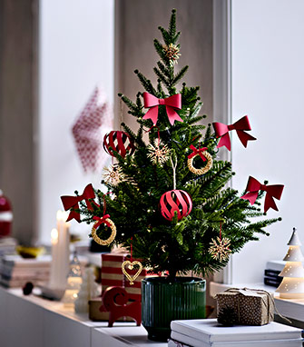 Pequeño árbol de Navidad en maceta verde sobre un alféizar, decorado con lazos y adornos de papel rojos y dorados.