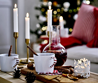 Primer plano de tazas blancas con asas de bastón de caramelo, vino caliente en jarra y especias en una mesa de madera.