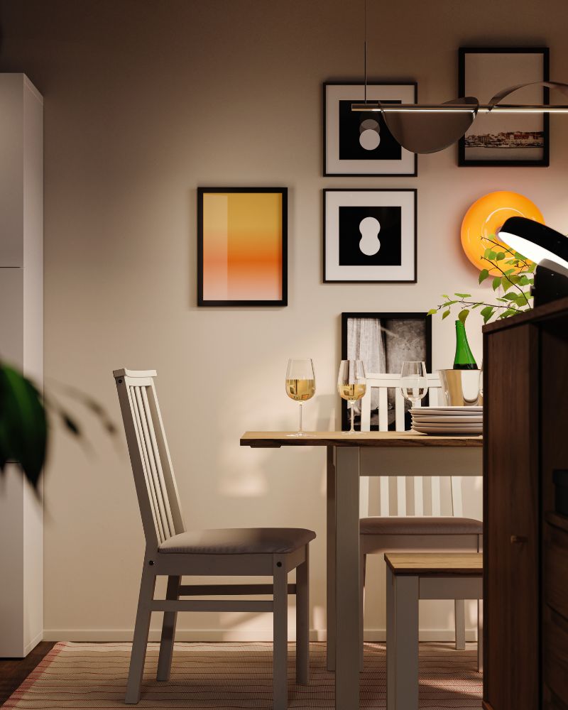 Un área de comedor de noche con un conjunto de comedor ÅLHULT que incluye una mesa extensible, un banco y sillas, y una lámpara LED redonda de color naranja.