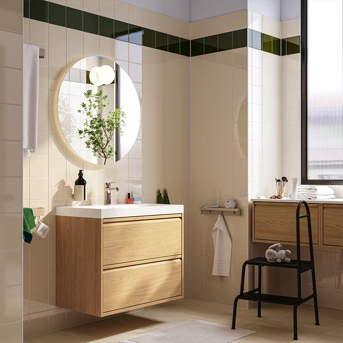 Modern bathroom with wall-mounted oak vanity, round mirror, and green tile accent border. Black step stool provides extra storage and functionality