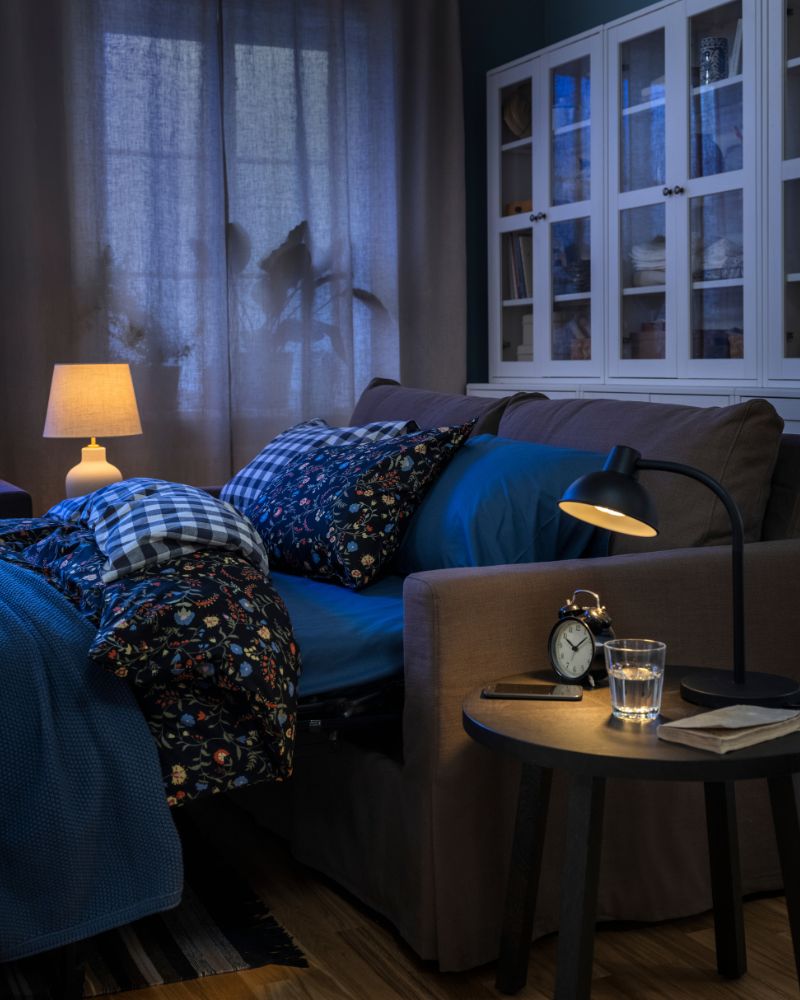 Un sofá cama HYLTARP, configurado como cama y con ropa de cama BRUDKRUSBÄR, se encuentra frente a varias combinaciones de muebles de almacenamiento HAVSTA.