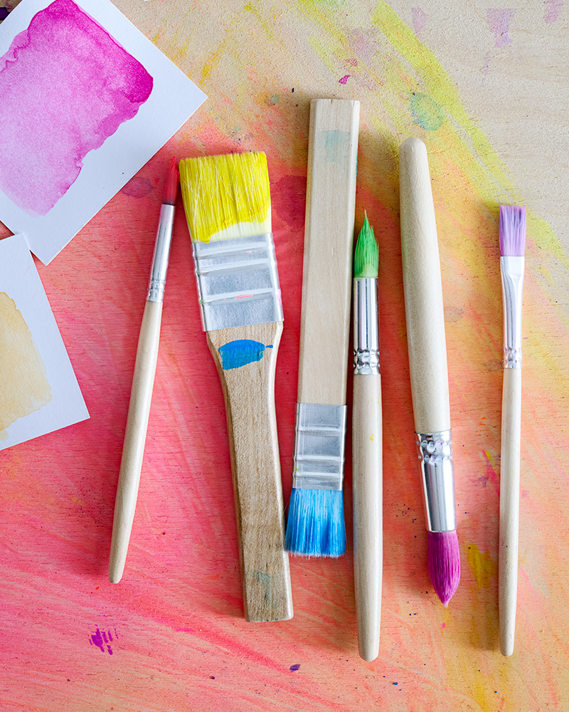 The 6 different sizes of MÅLA paint brushes, all dipped in different colours, laying on a wooden base.
