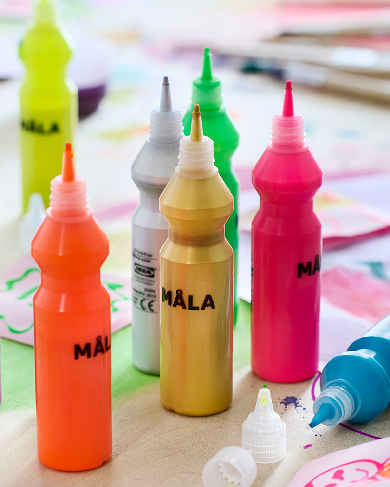 Close-up of 8 bottles of MÅLA fluorescent paint with lots of different paintings and drawings around them on the table.