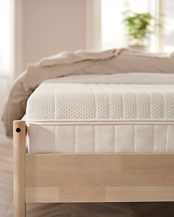 White mattress on natural wood bed frame in bright bedroom. Clean bedding and soft natural light create a calm sleeping space