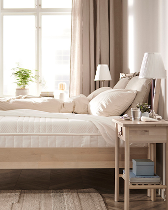 A bright bedroom with natural wood bed frame, beige bedding, and bedside table with lamp. Clean lines and warm neutral tones create a calm sleeping space