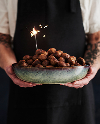 Persona con delantal negro sostiene cuenco cerámico lleno de trufas de chocolate, decorado con bengala encendida para celebrar