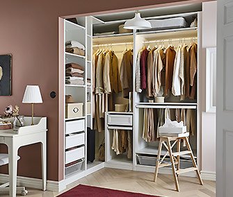 Armario abierto PAX modular en esquina, color blanco, con iluminación interior cálida, ropa colgando y cajones, en una habitación rosa pálida.