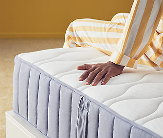 Mano toca un colchón de muelles embolsados blanco y gris con costuras laterales, una persona en pijama a rayas amarillas y blancas encima.