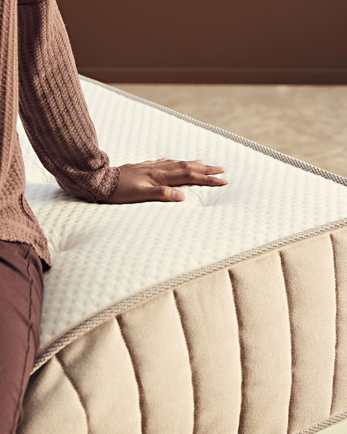 Mano toca la superficie acolchada de un colchón beige, mostrando el borde de tejido acolchado con costuras gruesas y beige.