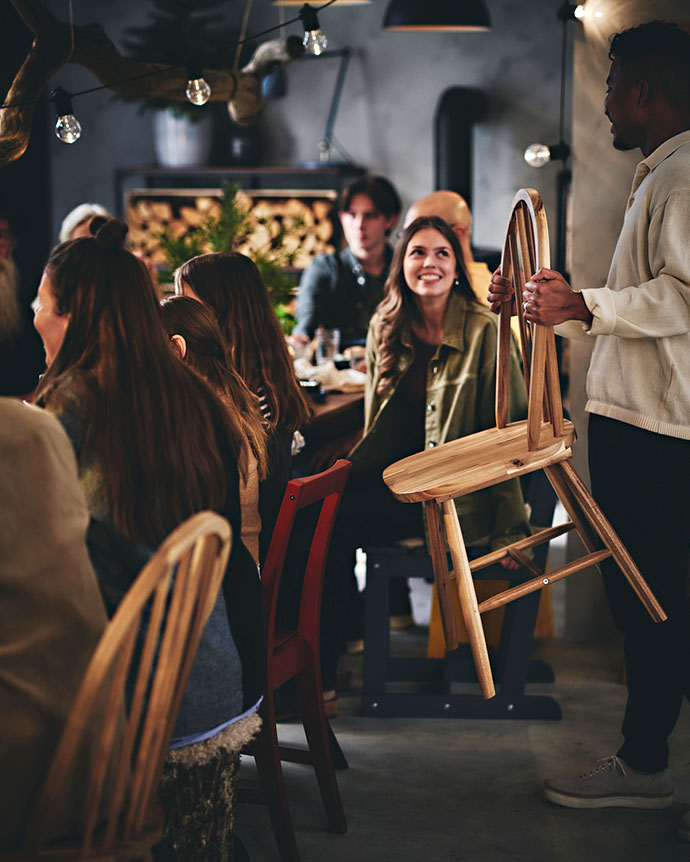 Un grupo de personas sentadas alrededor de una mesa rústica con sillas de madera, una de ellas siendo entregada por un hombre.