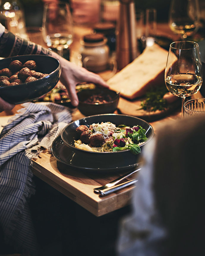 Una persona sirviendo un plato con albóndigas y ensalada en una mesa rústica con copas de vino, pan y especias, durante una cena.
