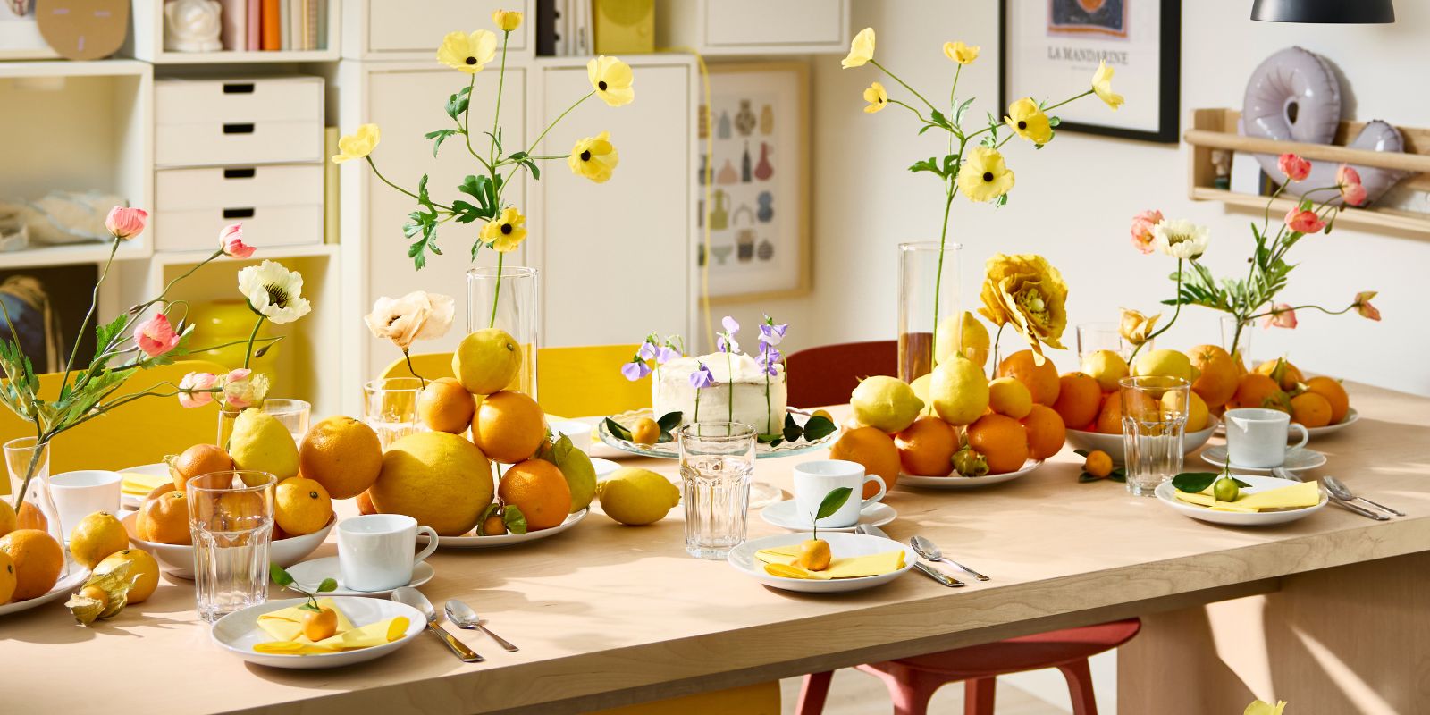 Una mesa BERGSHYTTAN tiene una decoración con platos blancos y vasos POKAL, además de naranjas y limones decorativos colocados sobre los platos.