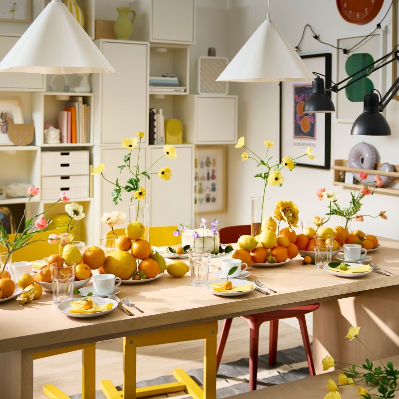 Una mesa BERGSHYTTAN tiene una decoración con platos blancos y vasos POKAL, además de naranjas y limones decorativos colocados sobre los platos.
