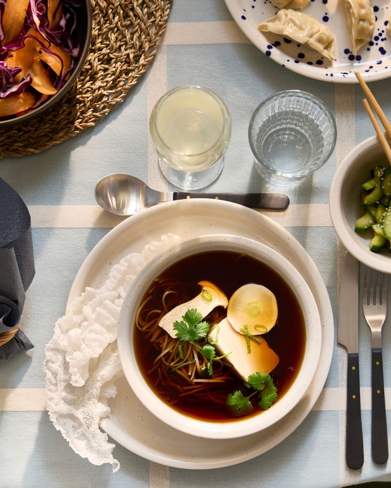 Un plato y un cuenco DAGGASTER de color beige claro con puntos contienen una sopa hecha de champiñones, tofu y fideos, junto a un cuenco con aguacates.