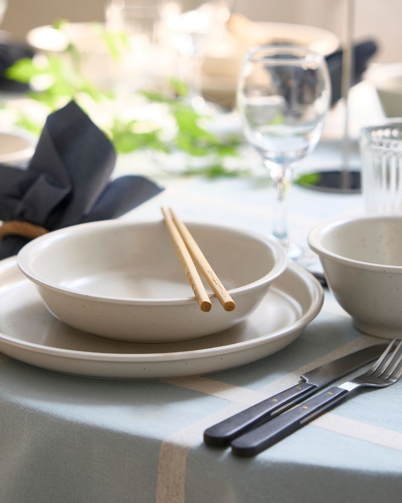 Un cuenco DAGGASTER beige claro con puntos está colocado sobre un plato, con dos palillos encima del cuenco y cubiertos a un lado.