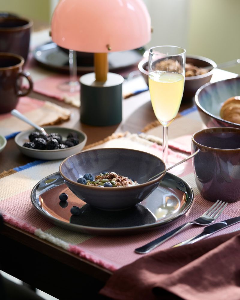 Un cuenco con yogur, muesli y arándanos está colocado junto a una taza GLADELIG de color lila y una copa de champán.