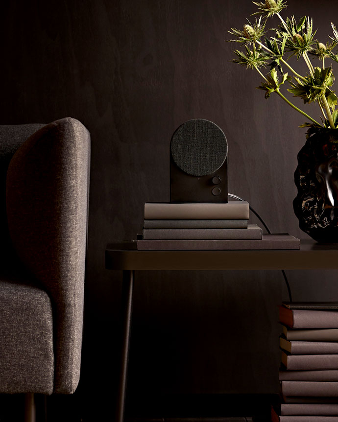 Un altavoz/dispositivo gris oscuro con tapa redonda de tela reposa sobre una pila de libros, junto a un sillón tapizado gris y un jarrón negro con flores secas.