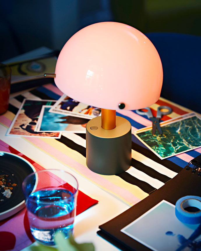 Lámpara portátil moderna en forma de hongo, con cúpula rosa y base gris mate, sobre tela de rayas colorida, fotos y artículos de escritorio.