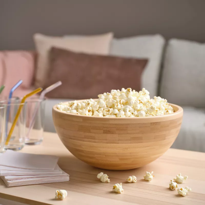 Bamboo bowl with popcorn and glasses with straws on the table, comfortable sofa in the background.