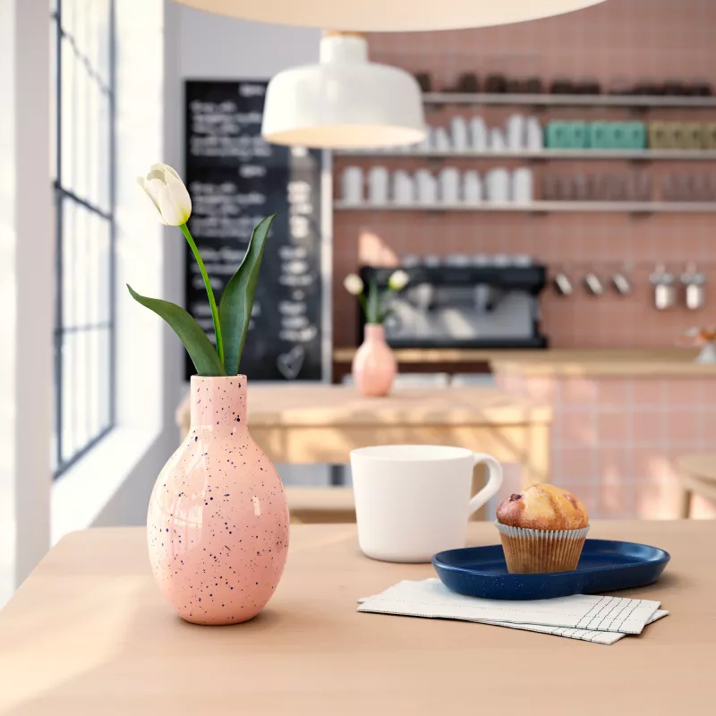 Coffee cup and cupcake close-up, with blurred vase and café setting in the background.