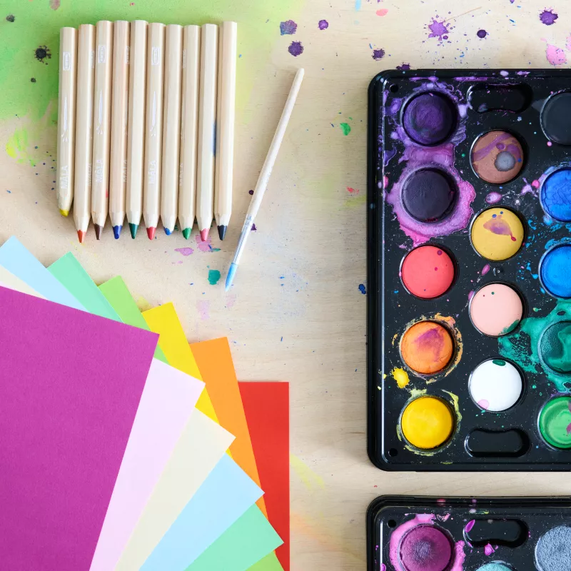 Colored papers, pencils, and watercolors seen from above with paint drops on the table.