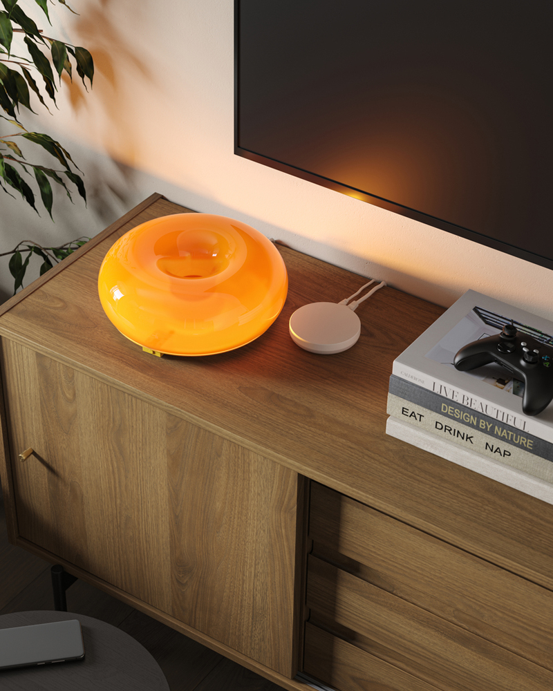 Una lámpara de mesa/pared LED VARMBLIXT se encuentra junto a un centro DIRIGERA y algunos libros sobre un mueble de TV RÅDMANSJÖ debajo de un televisor montado en la pared.
