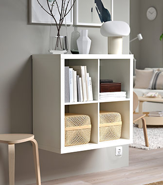 Estante KALLAX de 4 cubos blanco montado en pared con libros, cestos de ratán y una lámpara hongo encima.