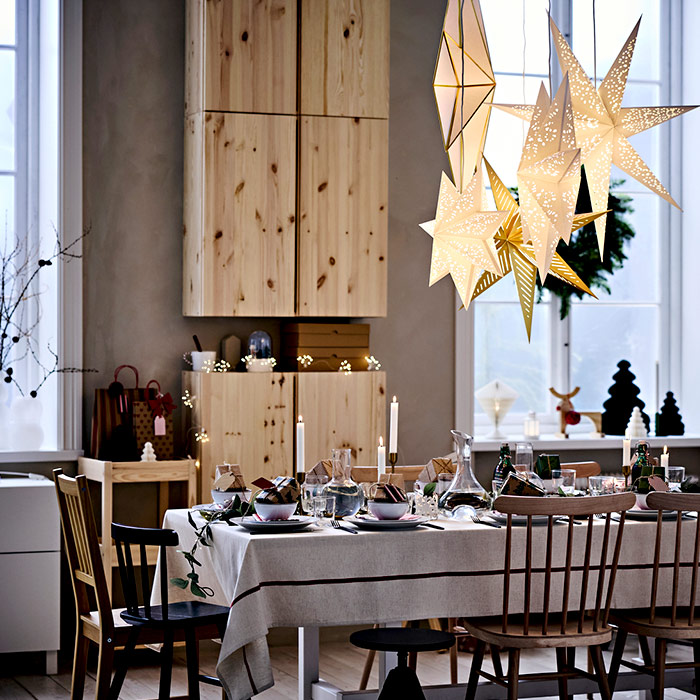 Mesa de comedor preparada para una cena navideña con mantel y decoración. Estrellas de papel iluminadas cuelgan del techo. Muebles de madera clara.