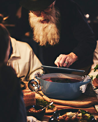 Close-up of an elderly man with a white beard serving a hot stew or soup from a blue cast iron pot on a wooden board.