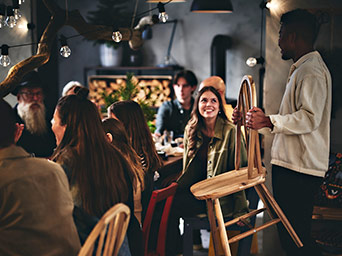 A standing man holding a wooden chair. Several people seated around a dining table, enjoying company and conversation.