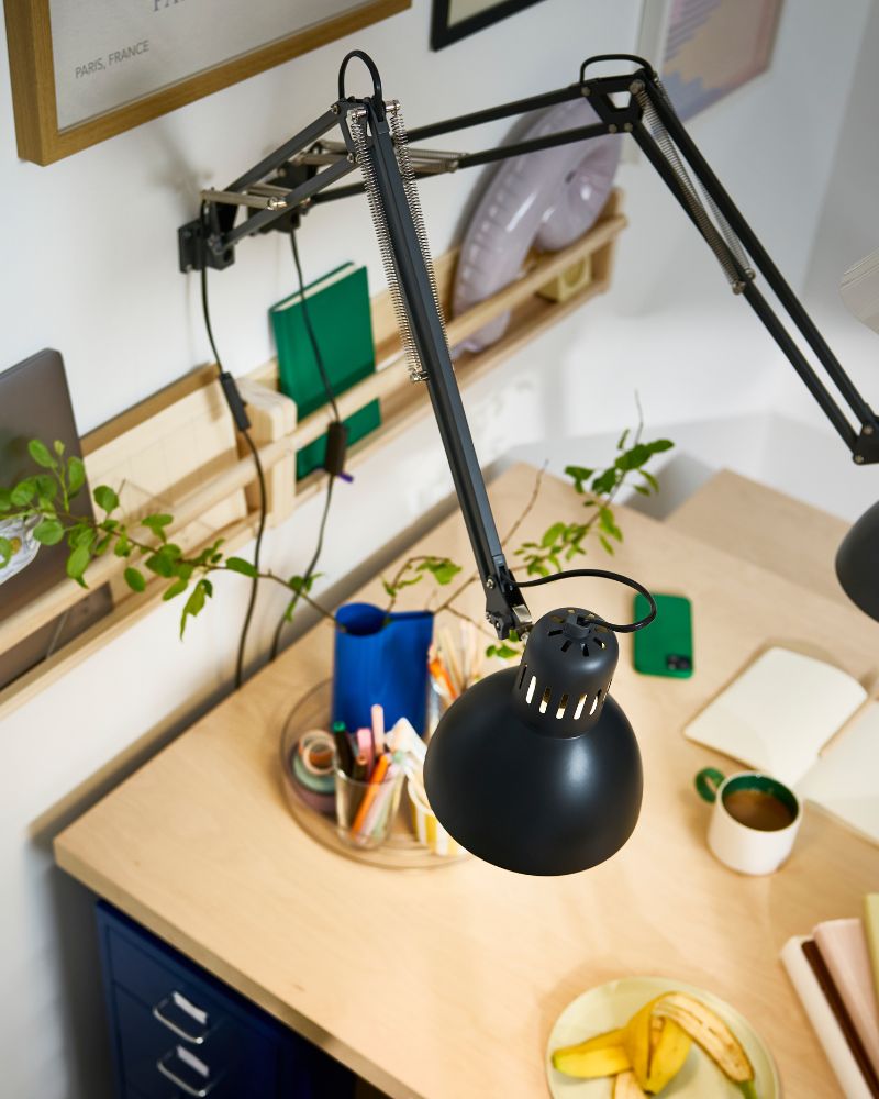 Two dark grey TERTIAL work lamps are attached to a wall with picture frames, shining their light over a BERGSHYTTAN table.