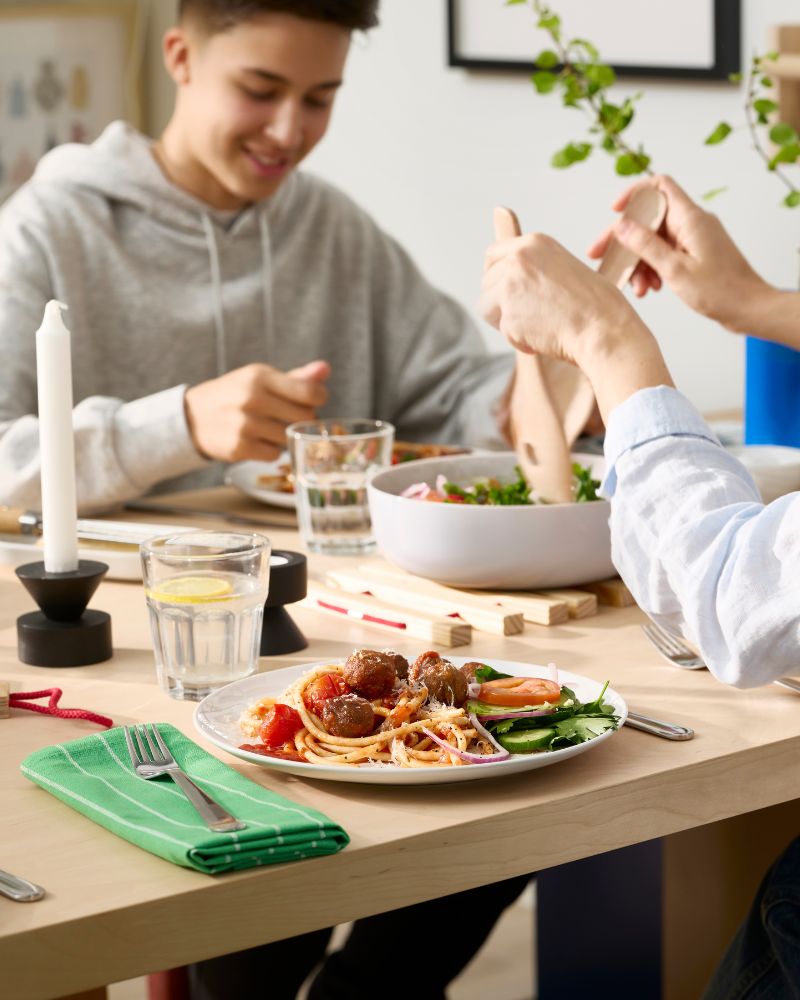 Two people are sitting at a dining table and eating food from white GODMIDDAG plates and drinking water from POKAL glasses.