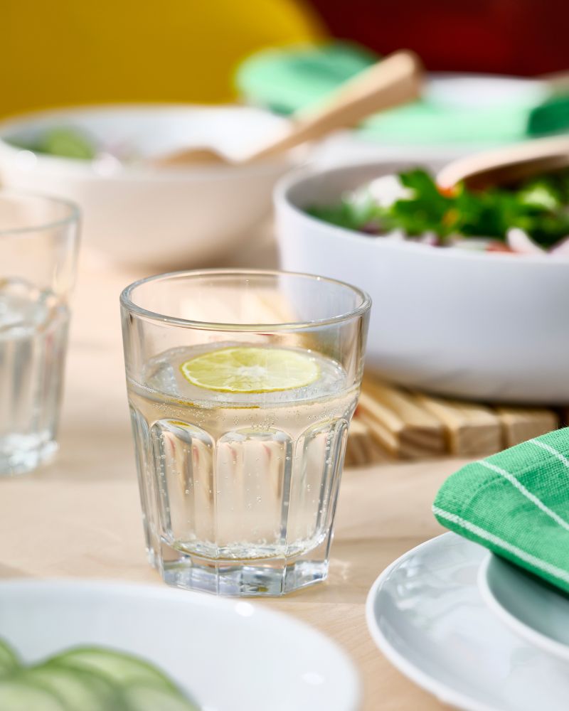 A POKAL glass with water and a slice of lemon is standing on a table filled with white plates and a VITBLECKA serving bowl.