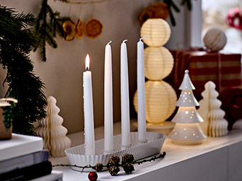 Cuatro velas blancas de Adviento en un candelabro acanalado, una encendida. Decoración navideña simple con faroles y un árbol de cerámica blanco.
