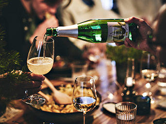 Pouring a sparkling drink from a green bottle into a wine glass, with food on the table and people dining in a warm setting.