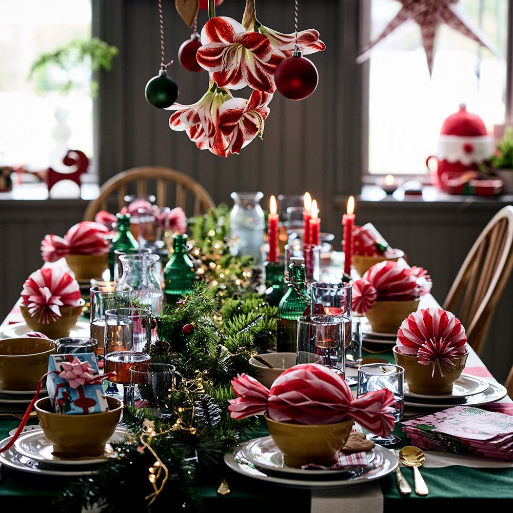 A Christmas table with VINTERFINT tree ornaments, VINTERFINT bottle with cork, VINTERFINT artificial mistletoe, SANDSKADDA bowl and SANDSKÄDDA plates, plus FÖRNUFT cutlery set and yellow SKOGSTA chairs.