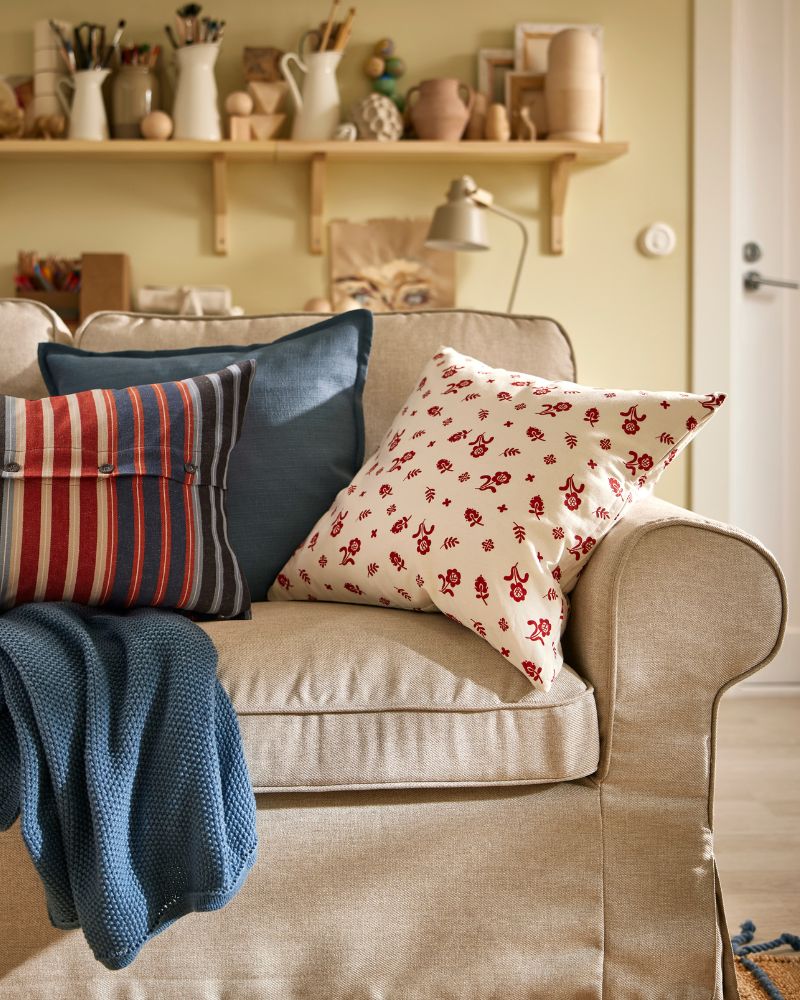 A blue-grey HUMLEMOTT throw and some cushions, including one with a floral TÅGSÄCKMAL cushion cover, lie on an EKTORP sofa.