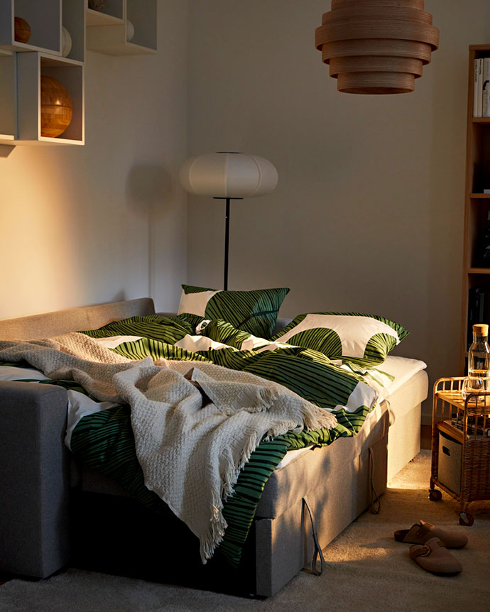 Cama convertible gris, abierta para dormir, con edredón de hojas verdes y una manta beige. Lámpara de pie y techo de madera.