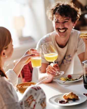 A smiling dad and his teenaged daughter sitting at the breakfast table clink their glasses of orange juice together.