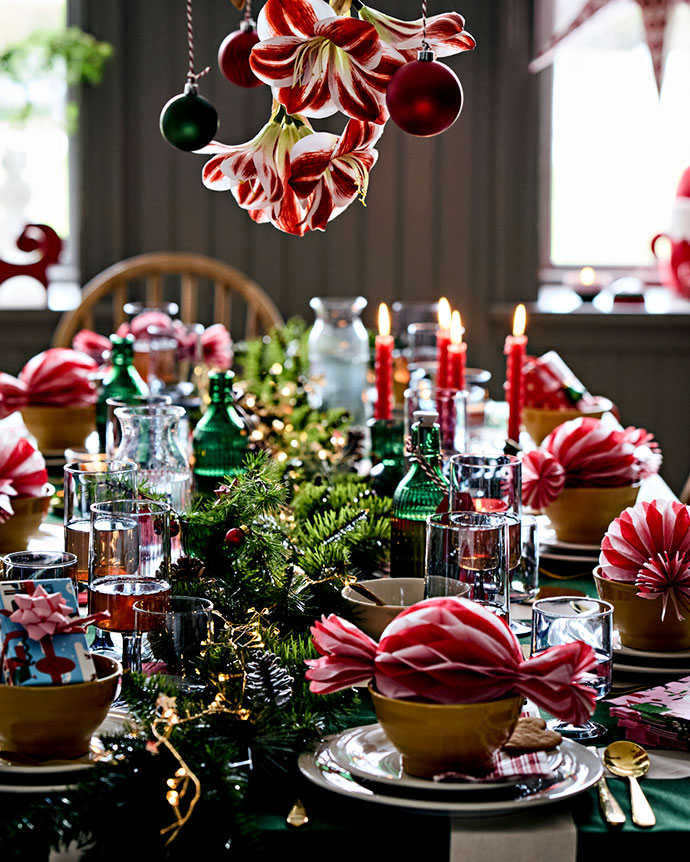 Christmas dining table with pine centerpiece and lights, green bottles, red candles, and hanging red flowers. Festive dinner setting.