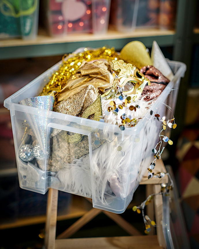 Clear plastic storage box filled with shiny gold and silver party supplies, like masks, tinsel, sequins, and white faux fur.