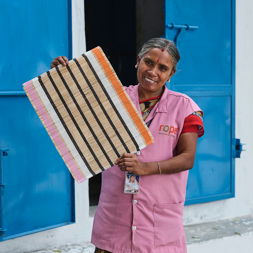 A person in pink clothes stands smiling in front of a pair of blue doors and holds up a striped MÄVINN place mat.