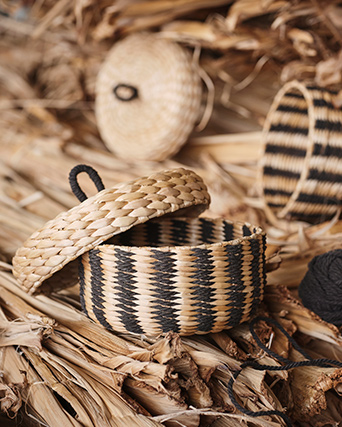 A black/beige striped MÄVINN box with lid sits on a pile of banana bark fibres. Next to it lies a ball of black cotton.