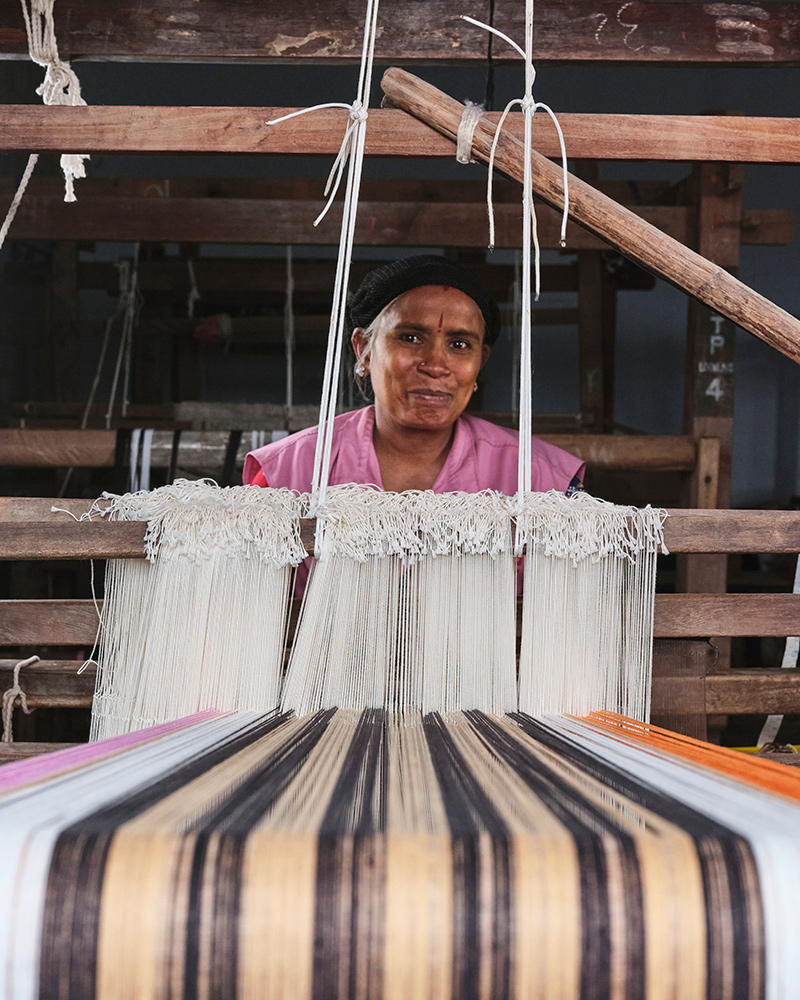 A person is sitting at a loom weaving a MÄVINN tablerunner out of banana bark fibres and cotton threads in vibrant colours.