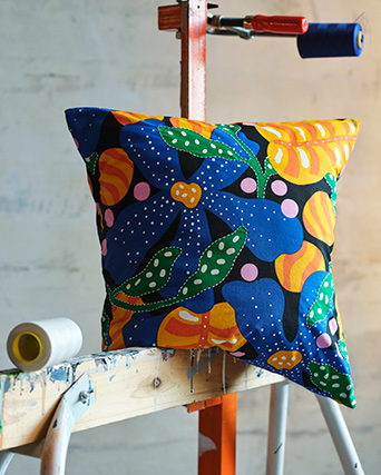 A MÄVINN cushion cover with a multicoloured floral pattern rests on a wooden beam stained with old paint.
