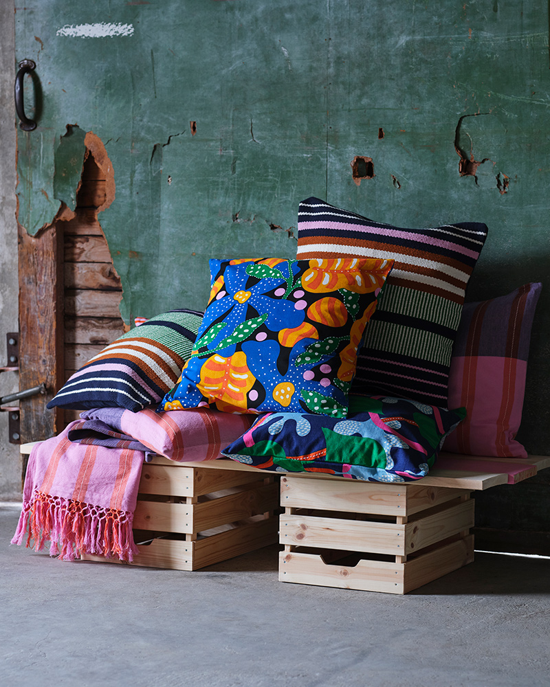 On top of a couple of wooden boxes are cushion covers and throws in pink, blue, green and orange from the MÄVINN collection