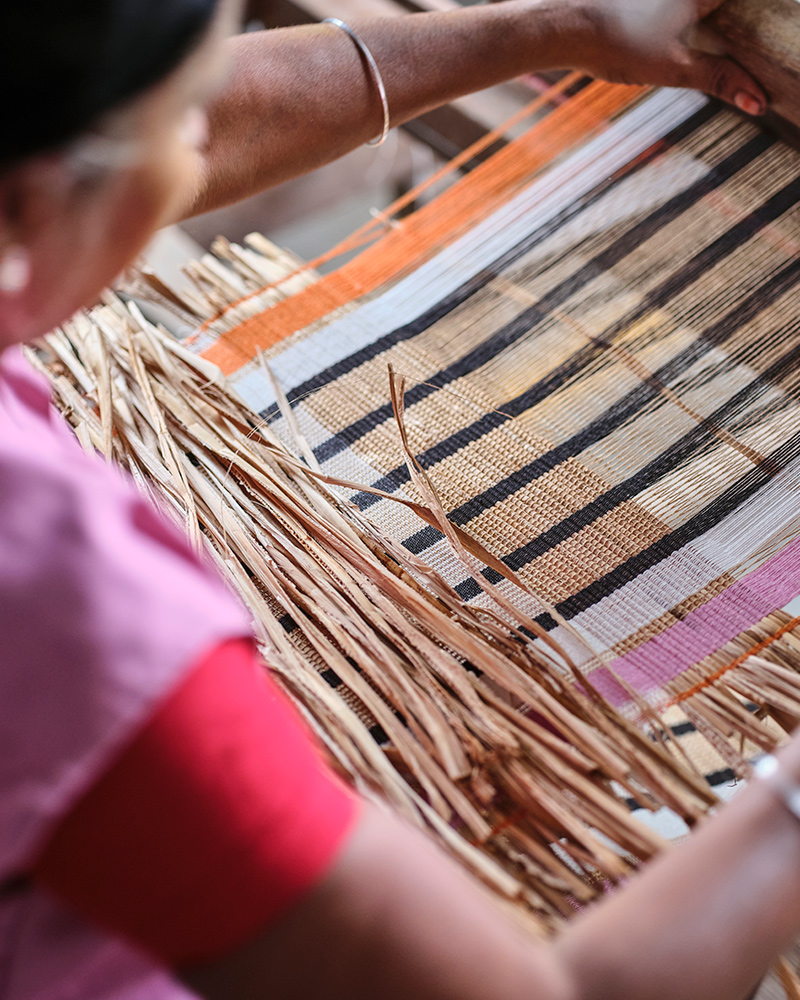 A person is weaving together banana fibres and cotton threads in orange, white, black and pink in a rustic weaving loom.