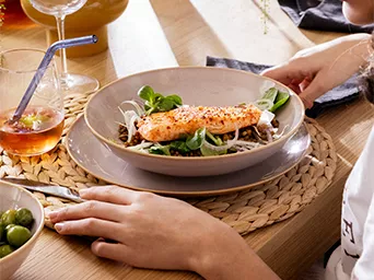 Manos sirviendo salmón con verduras en plato de cerámica, mesa de madera con mantel tejido y copa de vino